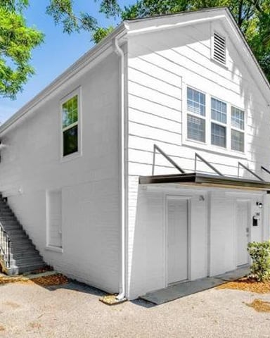White two-story house with garage below, metal stairs on left side, surrounded by green trees