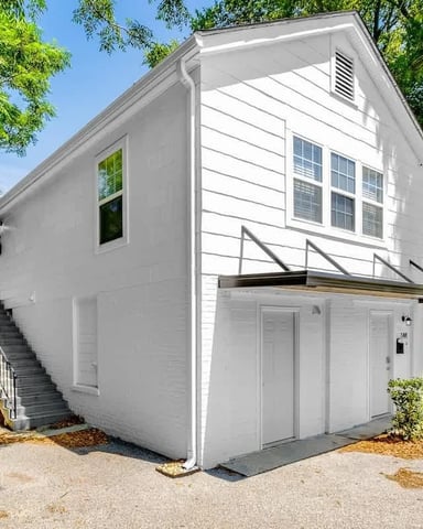 White two-story house with garage, metal staircase, and large windows surrounded by green trees