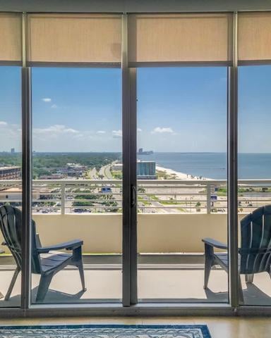 High-rise hotel room with floor-to-ceiling windows overlooking a beach, lake, and distant cityscape with two black Adirondack chairs on the balcony