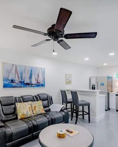 Open-plan living room with black leather reclining sofa, ceiling fan, and kitchen with stainless steel appliances