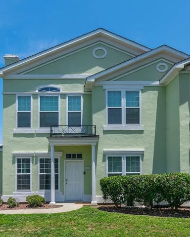 Two-story pale green house with white trim, gabled roof, and manicured landscaping under clear blue sky