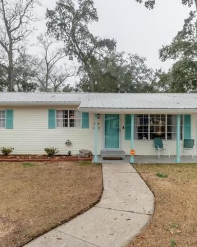 Single-story white cottage with turquoise shutters and porch, curved walkway, mature trees in background