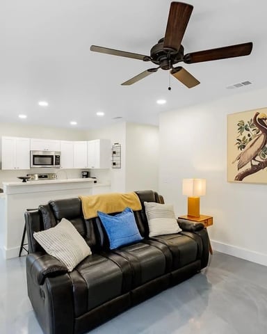 Open-plan living room with black leather sofa, white kitchen, ceiling fan, and recessed lighting