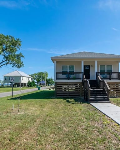 Elevated beach house with front porch and wooden stairs, surrounded by grass and neighboring homes under clear blue sky