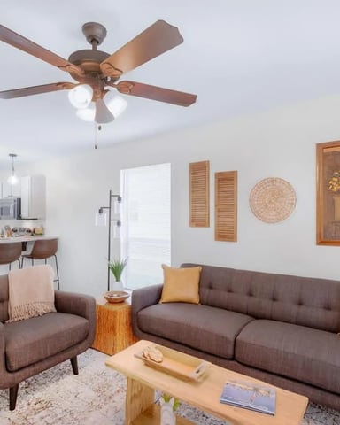 Open-plan living room with brown sofa, armchair, wooden coffee table, and ceiling fan with light fixtures overhead
