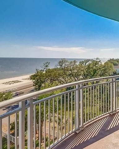 Elevated deck with metal railing overlooking sandy beach, vegetation, and calm water under blue sky