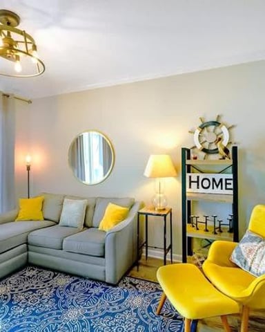 Bright living room with gray sectional sofa, yellow accent chair, blue patterned rug, and coastal decor including ship wheel and HOME sign.