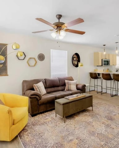 Open-plan living room with yellow armchair, brown sofa, kitchen with cream cabinets, and ceiling fan
