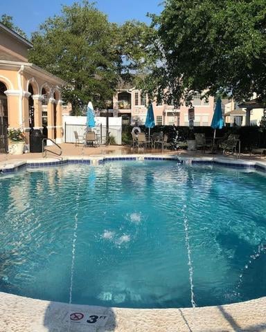 Resort swimming pool with blue umbrellas, lounge chairs, and peach-colored building with arched windows surrounded by trees
