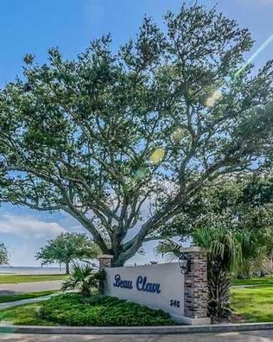Large oak tree with sprawling branches beside a brick sign reading "Beau Clair" at residential property entrance, waterfront view beyond