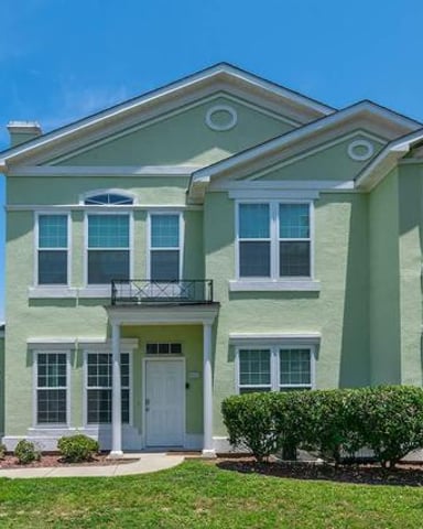 Two-story light green house with white trim, multiple windows, front porch, and manicured landscaping under clear blue sky