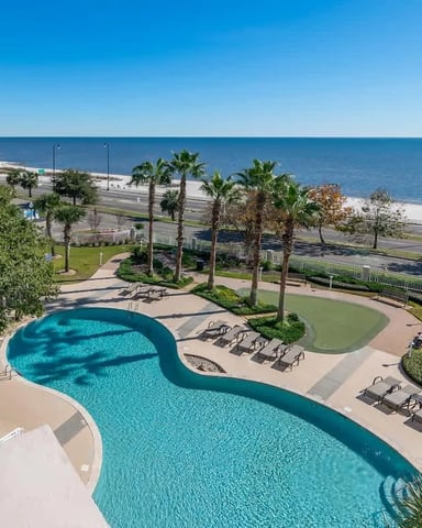 Curved resort pool with lounge chairs overlooking beach, ocean, and palm trees under clear blue sky
