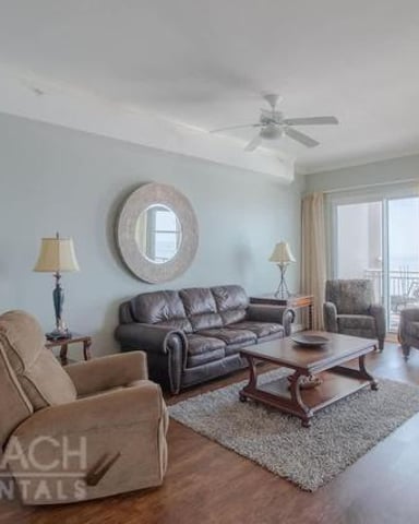 Spacious living room with brown leather sofa, beige armchairs, hardwood floors, and ocean view from balcony doors