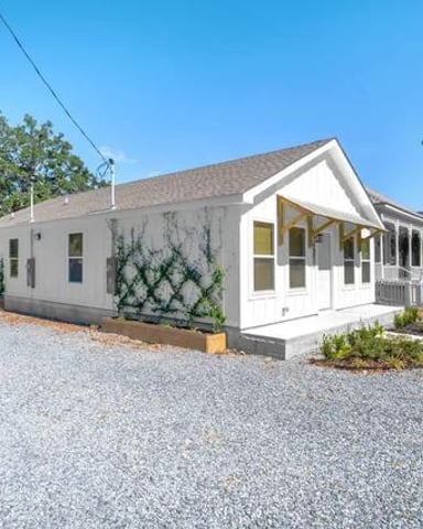 White cottage with front porch and gabled roof on gravel driveway surrounded by trees