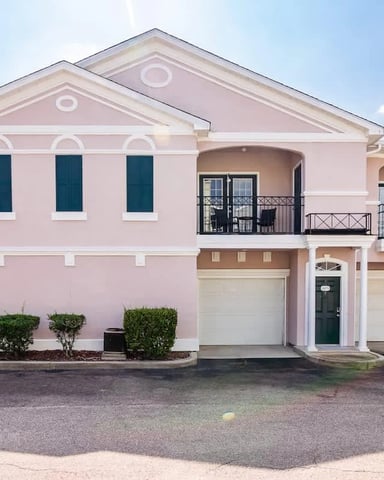 Pink two-story townhouse with dark blue shutters, balcony, and two-car garage in residential community