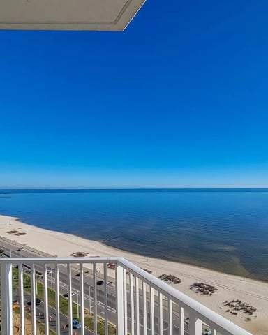 Balcony view of sandy beach, calm blue water, and distant shoreline with buildings under clear sky