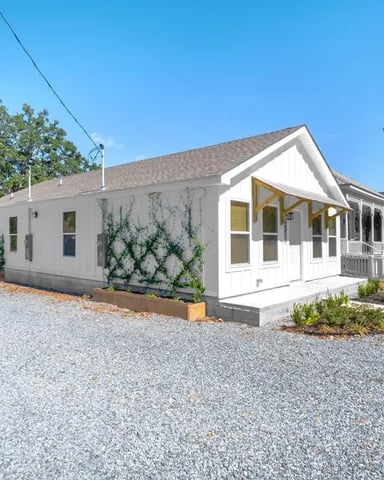 Modern white cottage with yellow-trimmed porch and gravel driveway, surrounded by mature trees and landscaping