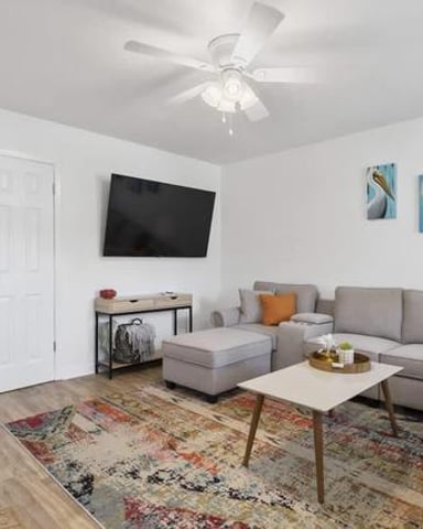 Bright living room with gray sectional sofa, wall-mounted TV, wooden coffee table, and bird artwork on white walls.