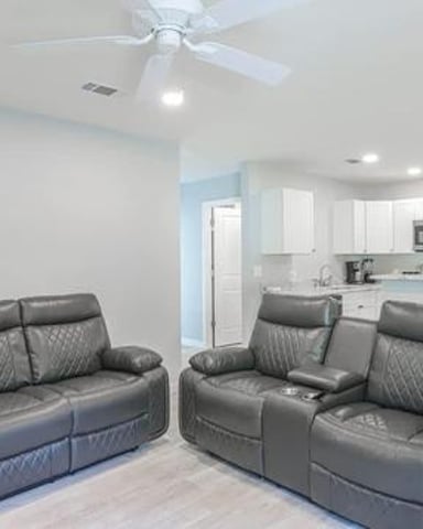 Open-plan living room with two gray leather recliners facing each other, ceiling fan, and kitchen with stainless steel appliances visible in background