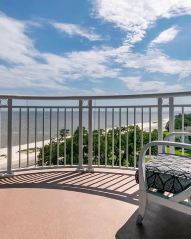 Curved balcony with metal railing, lounge chair, overlooking beach, river, and forested landscape under blue sky