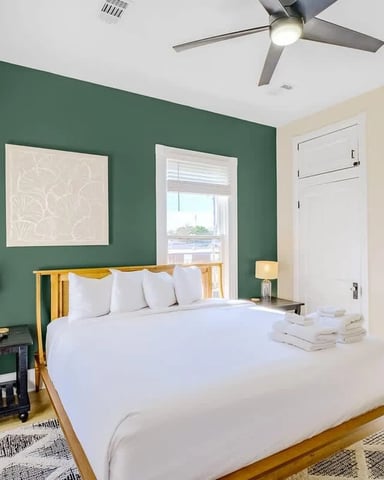 Bedroom with green accent wall, white bedding, wooden bed frame, nightstands with lamps, ceiling fan, and hardwood floors.