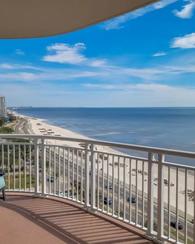 Beachfront balcony with turquoise chair overlooking sandy beach, ocean, and distant high-rise building under blue sky