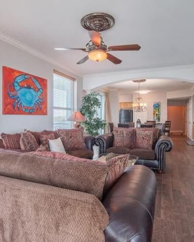 Spacious open-plan living room with beige sectional sofa, ceiling fan, large windows, and coastal artwork on walls