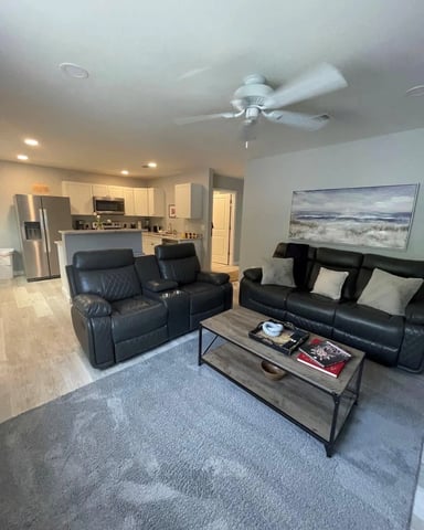 Open-plan living room with black leather recliners, coffee table, and kitchen with stainless steel appliances in background
