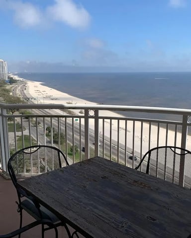 Elevated balcony with dining table overlooking sandy beach, coastal road, and ocean under blue sky