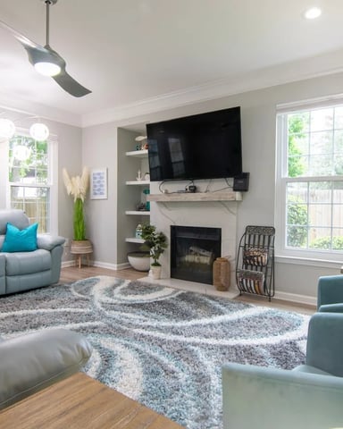 Bright living room with gray sectional sofa, fireplace with mounted TV, patterned area rug, and large windows overlooking greenery