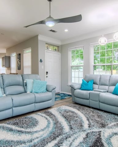 Bright living room with two light blue recliners, patterned area rug, large windows, ceiling fan, and pendant lights