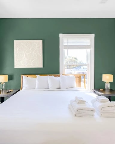 Bedroom with green accent wall, white bedding, wood headboard, side tables with lamps, and water view through glass doors