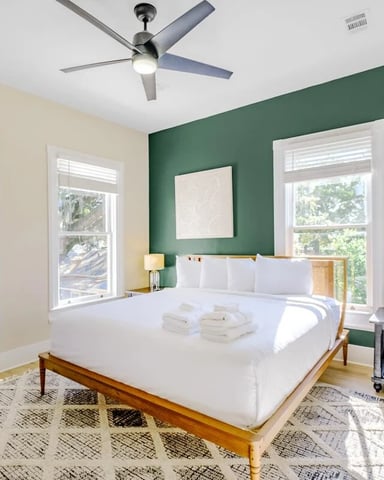 Bedroom with green accent wall, white bedding, wood bed frame, two windows, ceiling fan, and hardwood floors