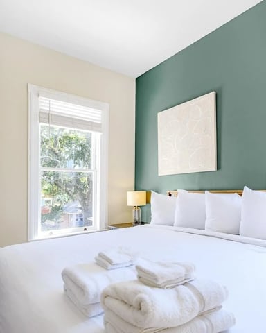Bedroom with white bed, folded towels, green accent wall, cream wall, window with garden view, and bedside lamp