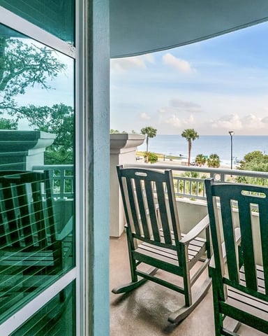 Oceanfront balcony with green rocking chairs overlooking beach, palm trees, and calm sea