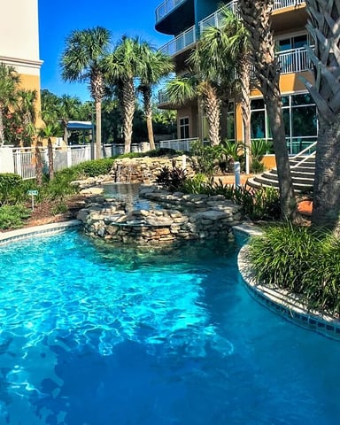 Resort pool with turquoise water, rock waterfall feature, palm trees, and colorful multi-story buildings surrounding courtyard