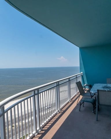 Oceanfront balcony with turquoise awning, dining table, chairs, and metal railing overlooking calm sea.