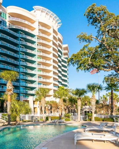 Beachfront resort with curved multi-story building, pool, palm trees, and lounge chairs under clear blue sky