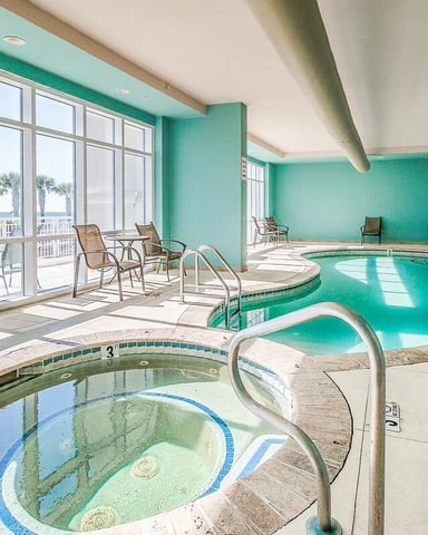 Indoor pool facility with hot tub, turquoise walls, large windows overlooking ocean and palm trees, lounge chairs, and exposed ceiling beams.