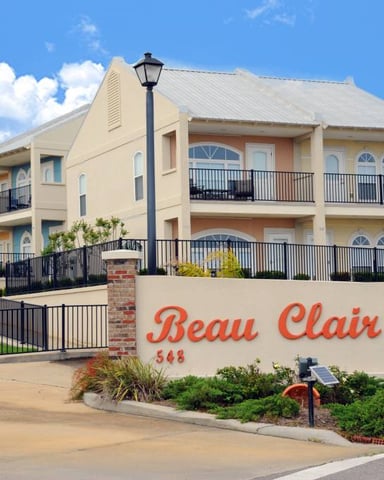 Multi-story beige residential building with black railings and brick pillars, entrance sign reading Beau Clair, clear blue sky