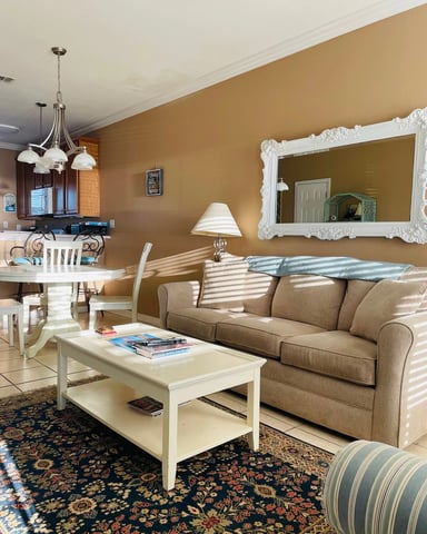 Open-plan living room with tan walls, beige sofa, white coffee table, ornate mirror, and kitchen visible in background