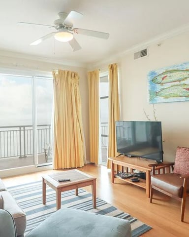 Bright living room with gray sofa, wooden furniture, TV, and glass doors opening to a balcony with ocean view