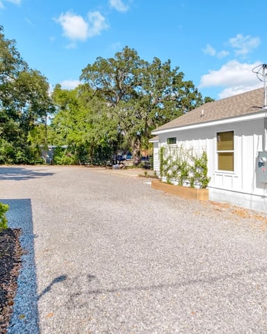 White single-story house with gravel driveway, landscaped planter beds, and large oak trees in background