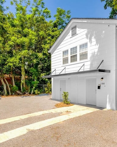 White modern house with two-car garage and gabled roof, surrounded by mature green trees and gravel driveway.