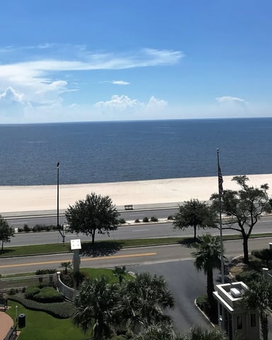 Beachfront view with sandy shore, calm ocean, coastal road, palm trees, and blue sky with white clouds