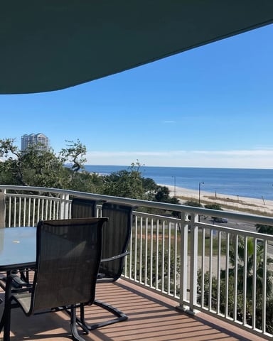 Elevated deck with dining table overlooking sandy beach, ocean, and distant high-rise building under clear blue sky