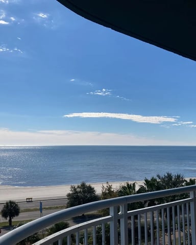 Oceanfront balcony view with white railing overlooking sandy beach, calm blue water, and clear sky