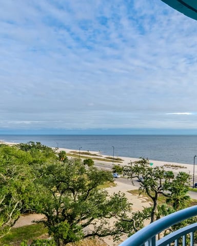 Elevated balcony view of sandy beach, calm ocean, coastal road, and high-rise building in distance