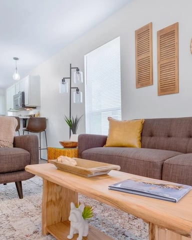 Modern living room with brown upholstered sofa and armchair, wooden coffee table, and open kitchen with white cabinets in background