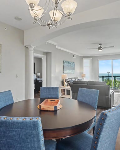 Open-plan dining and living room with round table, blue upholstered chairs, ocean view from balcony, and gray sofa